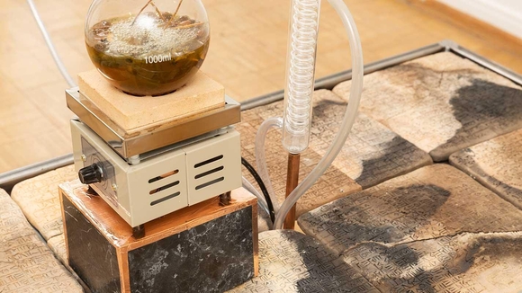 A mechanical contraption with a glass bowl, coiled glass tube, and metal body sits atop stone writings