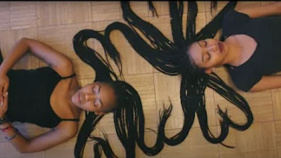 Two black women in black clothing, lying down with their braided hair placed in the shape of hearts.