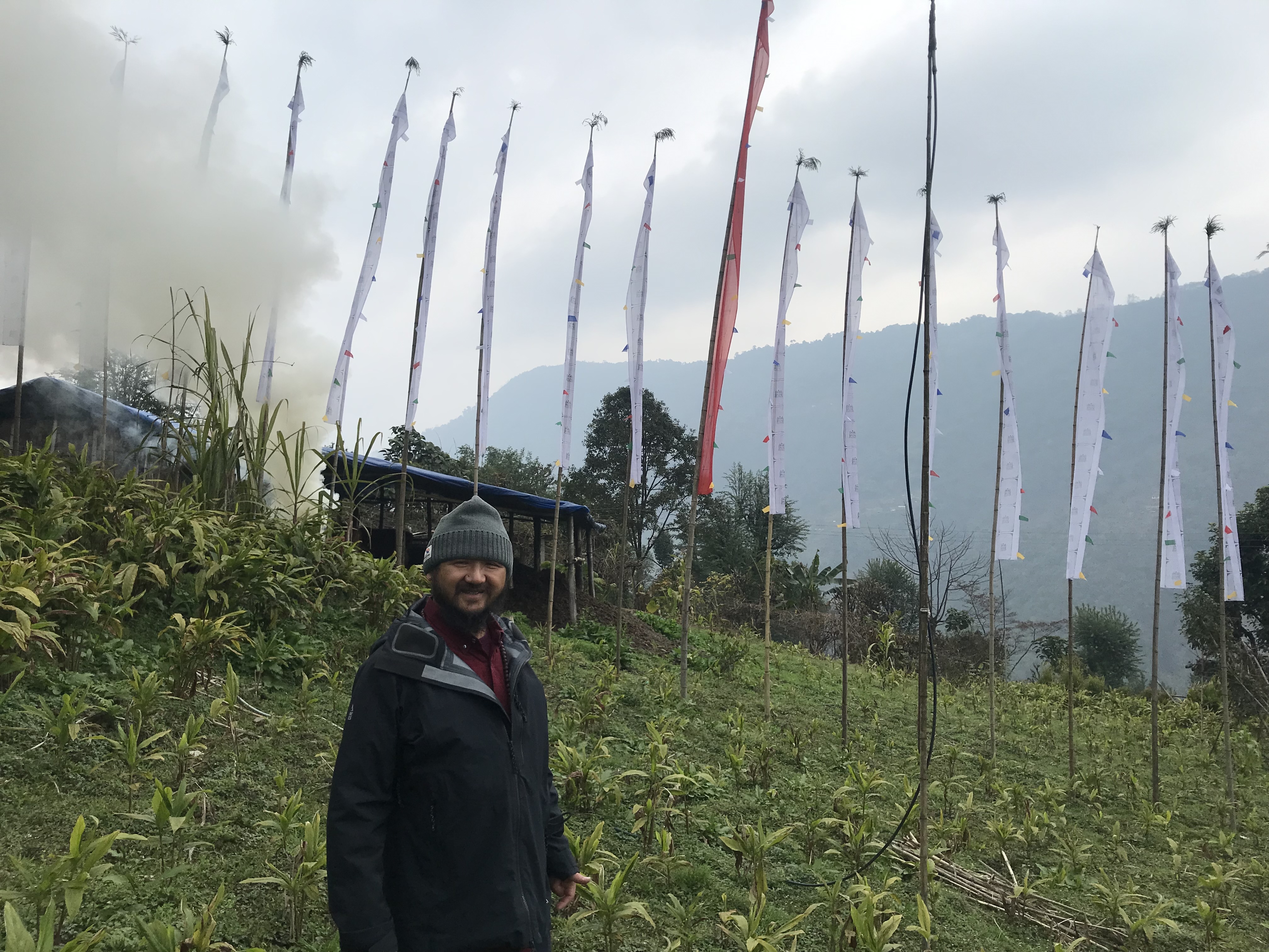 Photo of a person wearing a beanie and jacket, standing in front of a green hillside with flag poles behind them. 