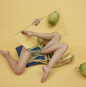 Two disembodied legs hidden by a mirror, surrounded by melons, on a yellow background