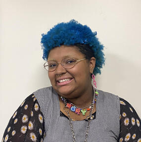 Portrait of Rory Hayes. They have blue hair and are smiling, looking into the camera, standing in front of a white background. 