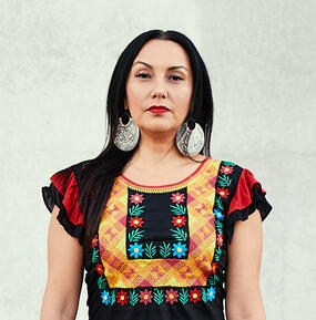 Felicia "Fe" Montes standing in front of a neutral background wearing a black shirt with colorful embroidery.