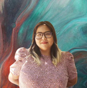 Portrait of Dria Yellowhair. Wearing a pink shirt and standing in front of a colorful background. 