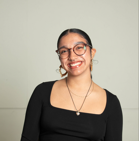 Portrait of a young person in glasses with their hair pulled back smiling into the camera. Wearing a black shirt and necklace. 