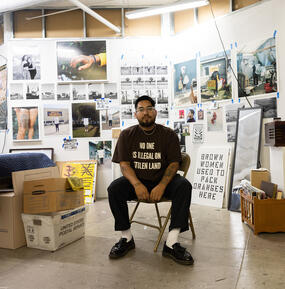Portrait of William Camargo sitting inside a studio space wearing a brown shirt. 