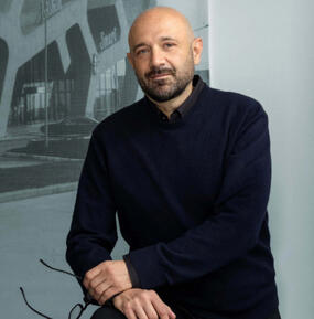 A bald person with facial hair wearing a black long sleeve shirt faces the viewer with his left hand gripping his left wrist. He looks content and serious.