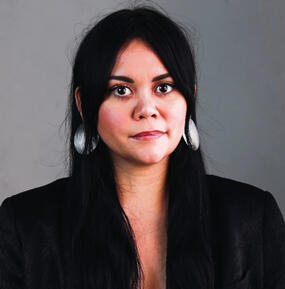 A person with long black hair and round silver earrings looks at the camera, serious.