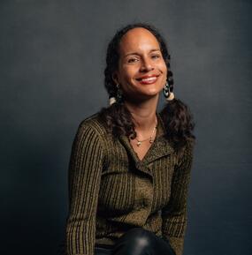 Portrait of Nina Collins. She is seated on a stool in front of a black background, smiling into the camera