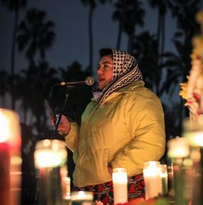 medium full-color portrait of Randa Jarrar speaking at a microphone, candles foreground
