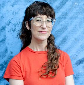 color closeup portrait of Alison O'Daniel wearing glasses and an orange t-shirt against a light blue background