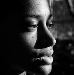 A shadow-cut profile portrait in black and white, one side of the face lit sharp and bright while the rest dissolves into darkness, the gaze lowered in quiet, unguarded thought.