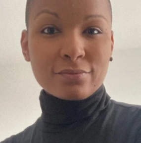 Close-up portrait of a Black person with a very short buzz cut, wearing a black turtleneck, looking directly at the camera with a slight smile against a plain light gray background.