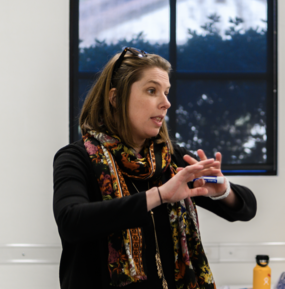 A brown-haired woman wearing a colorful scarf speaks to a classroom.
