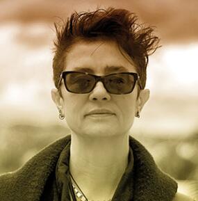 A sepia-toned portrait of a person with short, spiky reddish hair and dark sunglasses, wearing a dark coat, set against a blurred outdoor background.
