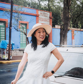 Kristina Wong posting in a white dress and sun hat in front of a blue building and palm trees/