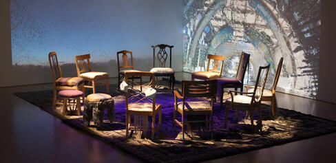 Cauleen Smith's installation of 13 chairs in a circle in dim lighting in front of a corner projection