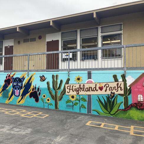 Completed mural at Yorkdale Elementary, showing a wide wall with a turquoise background, a sign that reads Highland Park, and a variety of painted symbols including sunflowers, a house, a panther, butterflies, and a girl reading a book. 