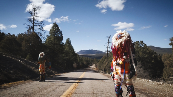 Two figures dressed in colorful, native-influenced clothing stand on a road with trees and mountains in the distance