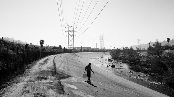 A black and white photograph of a man holding a bucket walking toward the LA River
