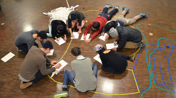 A group of people sit in a circle on the floor writing on sheets of paper. The floor is covered in shapes created by colored masking tape.