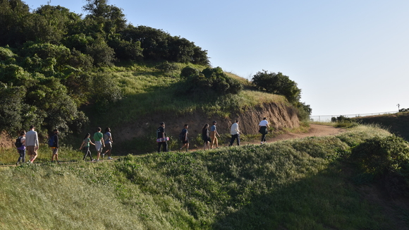 A wide image photograph of a group of people walking along a dirt path on a green hillside. 