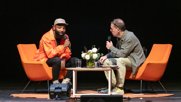 Stage view of Tavares Strachan and Paul Holdengräber sitting in orange chairs at angles across each other.