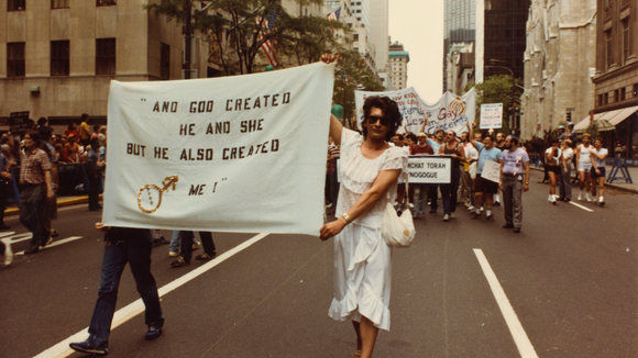 Archival image of Sylvia Rivera walking in a protest holding a large banner 