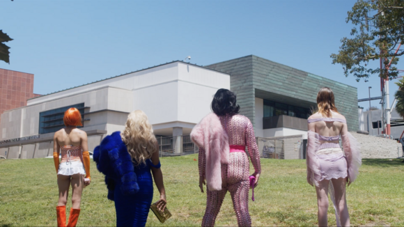 A still image of performers in vibrant, avant-garde outfits standing in front of a building. 