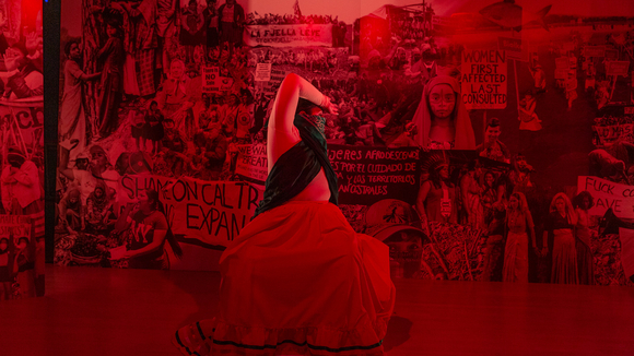 A woman lit in red bends backwards toward a wall covered in photos of people protesting