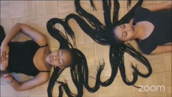 Two black women in black clothing, lying down with their braided hair placed in the shape of hearts.