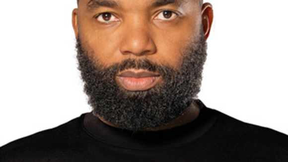 A man wearing a black turtleneck stares at the camera, with a serious expression against a white background