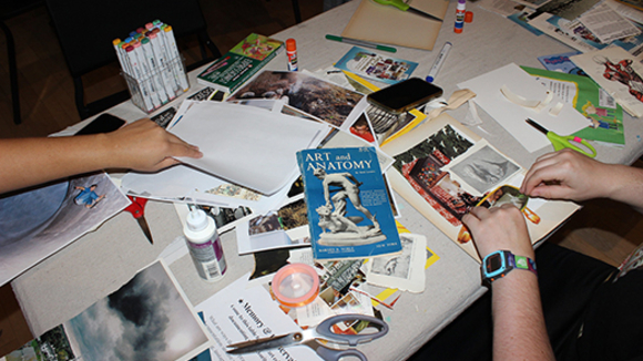 Magazines, scissors, glue and other materials on a table with hands creating a collage