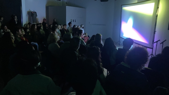 A group of people is seated watching a screening in the OXY ARTS gallery. The room is dark and the screen is lit with bright abstract colors. 