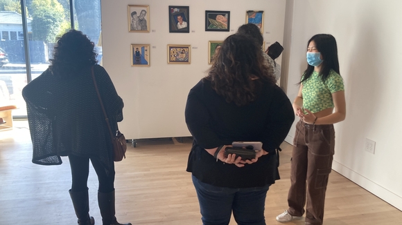 Photo of three people standing in an art gallery looking at paintings hung on the wall, their backs are turned away from the camera. 