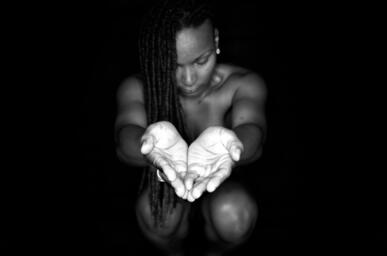 A black-and-white portrait showing a figure kneeling in darkness with long braided hair, gazing down at their own cupped hands extended toward the viewer and illuminated by a single light source against an otherwise black background.