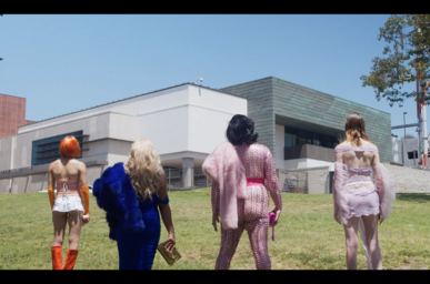 A still image of performers in vibrant, avant-garde outfits standing in front of a building. 