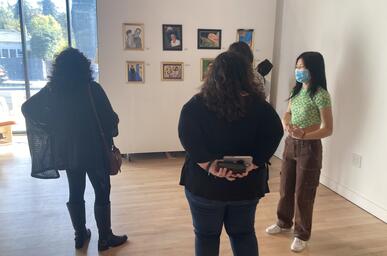 Photo of three people standing in an art gallery looking at paintings hung on the wall, their backs are turned away from the camera. 