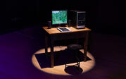 A desk stands in a spotlight surrounded by darkness, topped with a computer and monitor