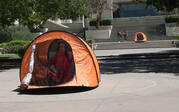 performer sitting in a small orange camping tent in the middle of the quad