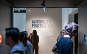 Close up of exhibition's title label on the wall, with people in foreground standing in hallway