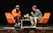 Stage view of Tavares Strachan and Paul Holdengräber sitting in orange chairs at angles across each other.