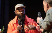 Close up of Tavares Strachan sitting and gesturing upwards as he speaks into a microphone to Paul Holdengräber.