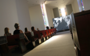 An angled view down the aisle of Herrick chapel, showing the audience in the pews leading up to a black and white frame from Last Whispers on a large screen.