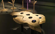 View of Jerome Dent's black glass hand-sized sculptures, in abstract stone-like forms on a wooden pedestal. Corn Stalk sculptures and projected video art is in background.