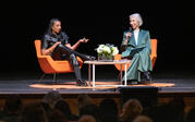 Full view of Robin Coste Lewis and Alexandra Grant sitting on stage. Lewis gestures and speaks with a furrowed brow while Grant listens.