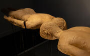 Close up of "Horizon." Two full-body, beige-clay ceramic casts levitate mounted on steel rods. They lie face down, their bottom layers missing, heads next to each other. 