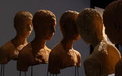 Roksana's ceramic work "Land" from behind, backlit in dramatic shadow. Five casted unglazed clay busts of the artist levitate at eye level in profile, mounted on steel rods.