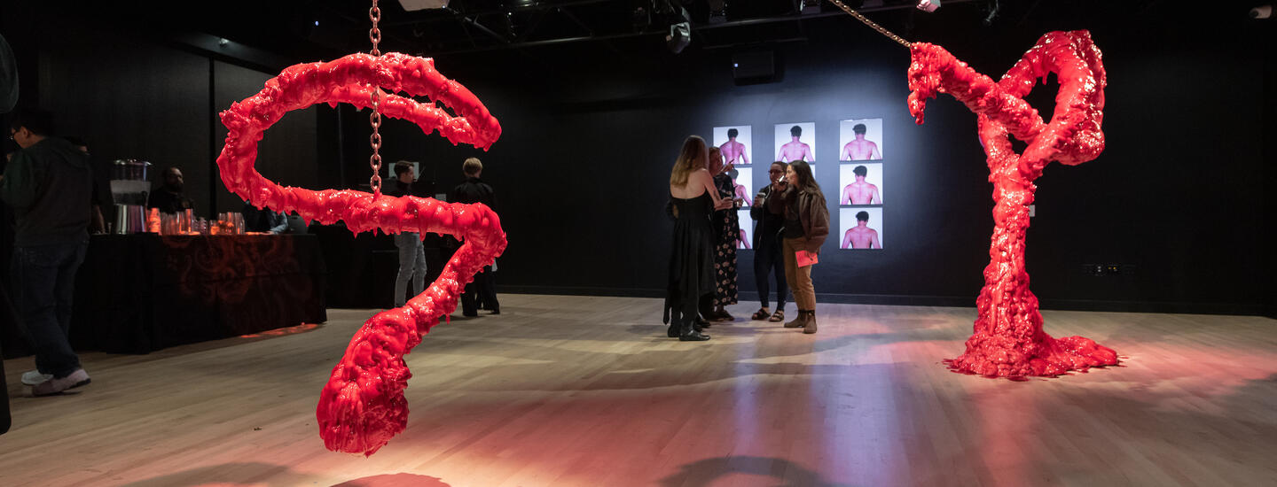 Two large, bulbous, red sculptures are suspended from the ceiling, with a video installation of nine shots of human backs in the background