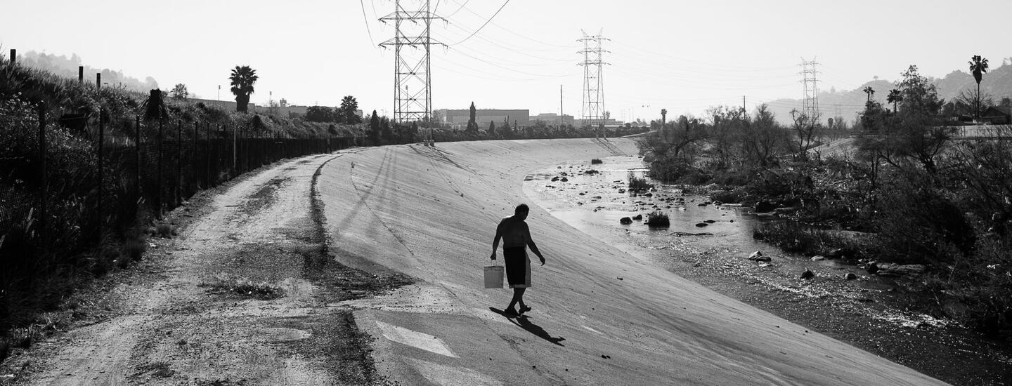 A black and white photograph of a man holding a bucket walking toward the LA River