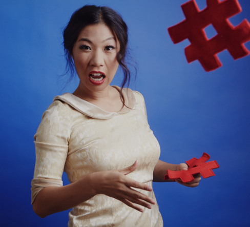 Portrait of Kristina Wong in a white dress standing in front of a bright blue background. She is throwing a red sculptural shape towards the camera. 